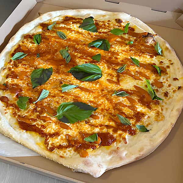 A whole round pizza with white cheese, orange sauce drizzle, and fresh basil leaves in a box.
