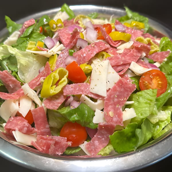 A vibrant Italian salad with salami, cheese, lettuce, tomatoes, onions, and peppers in a metal bowl.