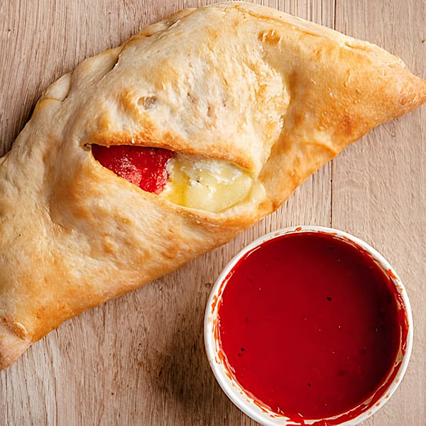 A golden-brown calzone with visible red sauce and melted cheese, next to a cup of red dipping sauce.